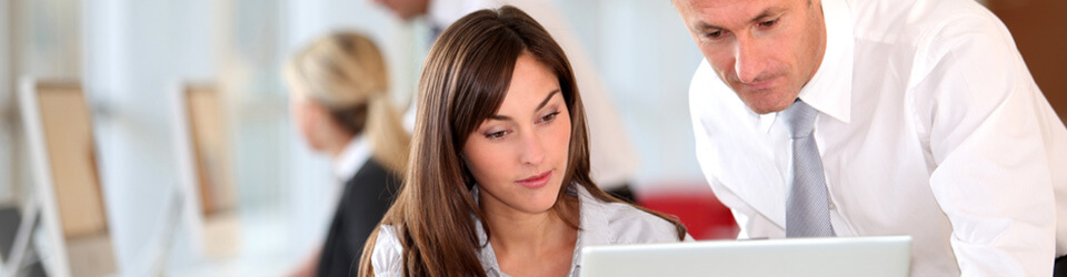 A businessman in a white shirt looks over the shoulder of a female colleague as she works on a white laptop in a minimalist, modern workspace.