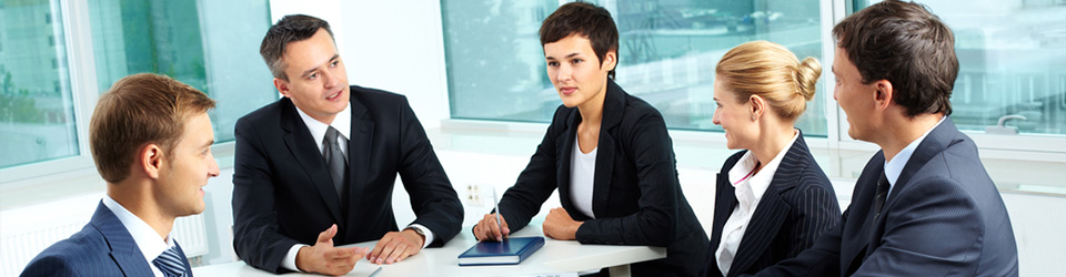 A group of businessmen and businesswomen take part in a meeting at a white table in an empty, white conference room.
